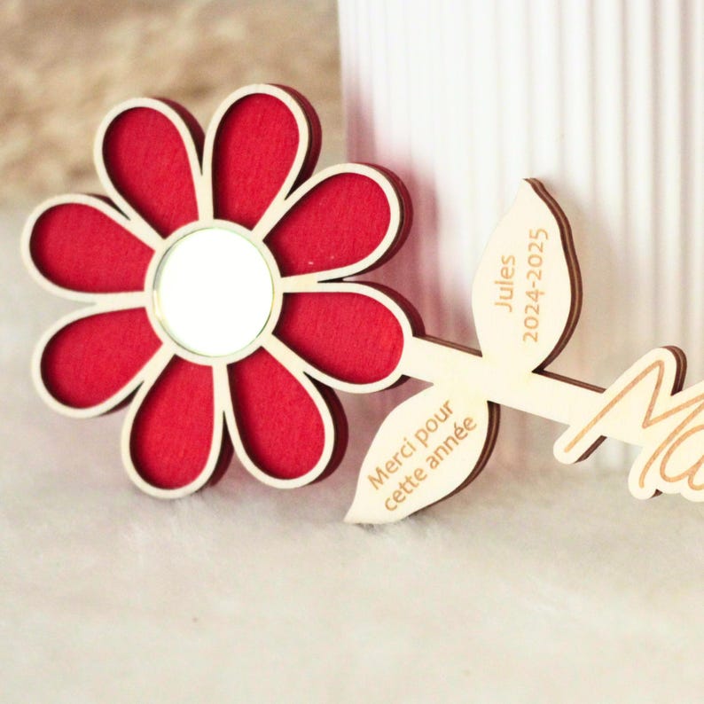 May include: A wooden flower-shaped decoration with a red center and a white border. The flower has a mirror in the center and is attached to a wooden stem with the text "Jules 2024-2025" and "Merci pour cette ann&eacute;e".