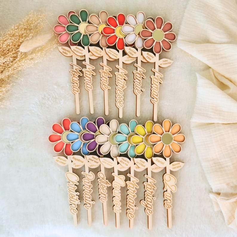 May include: Wooden flower-shaped toppers with colorful petals and a central disc. Each topper has a wooden stem with a name or word, such as "Mama" or "Mariage". The toppers are designed for various occasions.