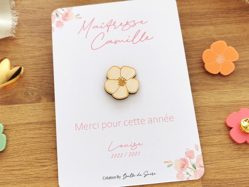 May include: A wooden flower pin on a white card with the text "Ma&icirc;tresse Camille" and "Merci pour cette ann&eacute;e". The card also includes the name "Louise" and the year "2022/2023". Decorative floral elements are present.