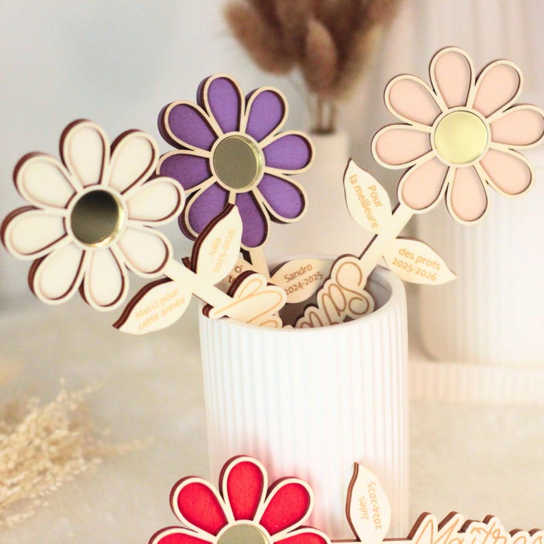 May include: Wooden flower-shaped gifts in various colors, including white, purple, pink, and red, with gold-colored centers. Each flower has a leaf-shaped stem with text. The flowers are displayed in a white ceramic vase.