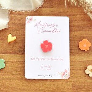 May include: A white card with the text "Ma&icirc;tresse Camille" and "Merci pour cette ann&eacute;e" in pink script, with a coral flower pin. Additional flower pins in orange, green, and cream are scattered around.