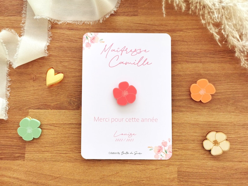 May include: A white card with the text "Ma&icirc;tresse Camille" and "Merci pour cette ann&eacute;e" in pink script, with a coral flower pin. Additional flower pins in orange, green, and cream are scattered around.