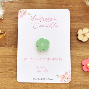 May include: A white card with pink floral accents and text in French, "Ma&icirc;tresse Camille" and "Merci pour cette ann&eacute;e". A light green flower pin is attached. Additional flower pins in pink and beige are also visible.