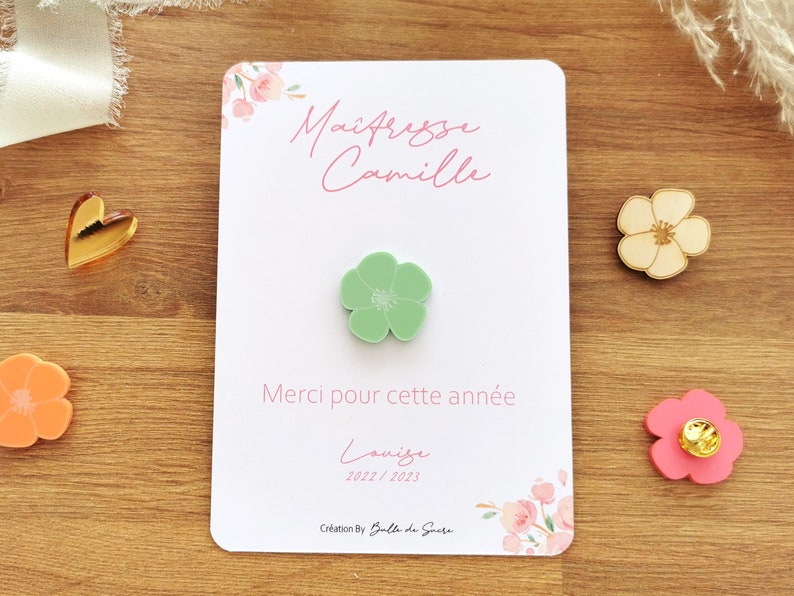 May include: A white card with pink floral accents and text in French, "Ma&icirc;tresse Camille" and "Merci pour cette ann&eacute;e". A light green flower pin is attached. Additional flower pins in pink and beige are also visible.