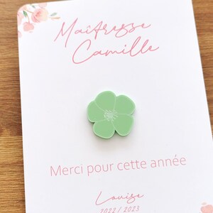 May include: A light green flower-shaped pin sits on a white card with pink floral accents. The card has the text "Ma&icirc;tresse Camille" and "Merci pour cette ann&eacute;e" in pink script. The name "Louise" and the year "2022/2023" are also visible.