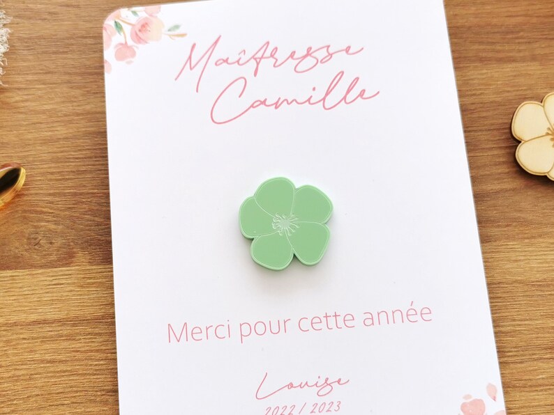May include: A light green flower-shaped pin sits on a white card with pink floral accents. The card has the text "Ma&icirc;tresse Camille" and "Merci pour cette ann&eacute;e" in pink script. The name "Louise" and the year "2022/2023" are also visible.