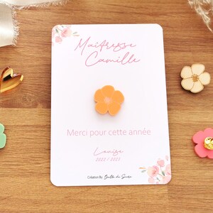 May include: An orange flower pin is centered on a white card with the text "Ma&icirc;tresse Camille" and "Merci pour cette ann&eacute;e". Additional flower pins in green, pink, and wood are scattered around the card. A gold heart is also present.