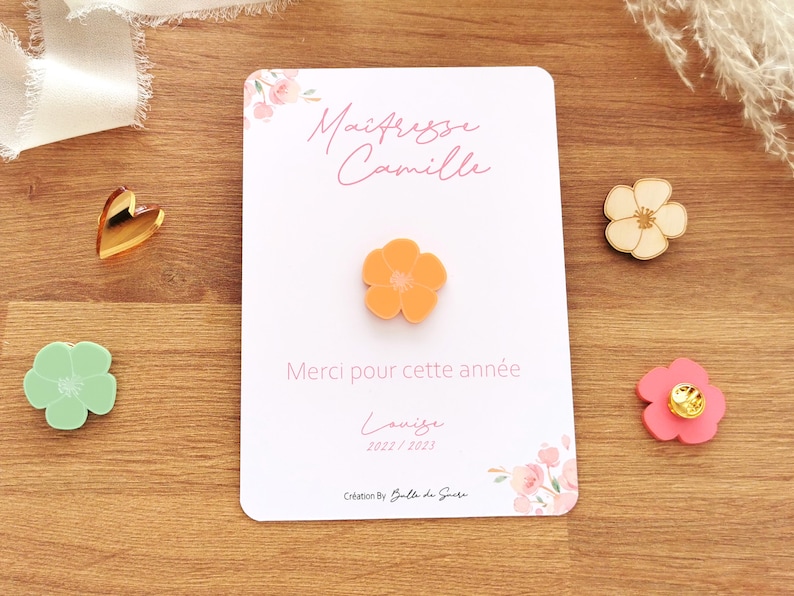May include: An orange flower pin is centered on a white card with the text "Ma&icirc;tresse Camille" and "Merci pour cette ann&eacute;e". Additional flower pins in green, pink, and wood are scattered around the card. A gold heart is also present.