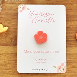 May include: A white card with the text "Ma&icirc;tresse Camille" and "Merci pour cette ann&eacute;e" in pink script. A coral-colored flower-shaped pin is centered on the card. Floral accents and the year 2022/2023 are also visible.