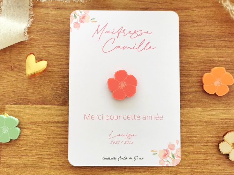 May include: A white card with the text "Ma&icirc;tresse Camille" and "Merci pour cette ann&eacute;e" in pink script. A coral-colored flower-shaped pin is centered on the card. Floral accents and the year 2022/2023 are also visible.
