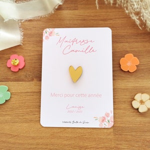 May include: A white card with pink floral accents and text in French, "Ma&icirc;tresse Camille" and "Merci pour cette ann&eacute;e." A gold heart-shaped pin is centered on the card. Decorative flower-shaped pins in pink, orange, and green are scattered around the card.