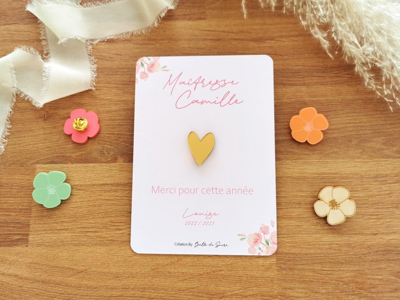 May include: A white card with pink floral accents and text in French, "Ma&icirc;tresse Camille" and "Merci pour cette ann&eacute;e." A gold heart-shaped pin is centered on the card. Decorative flower-shaped pins in pink, orange, and green are scattered around the card.