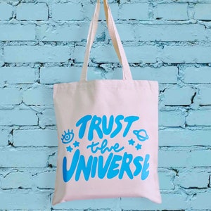 May include: A white canvas tote bag with blue text that says "TRUST the UNIVERSE". The text is in a hand-drawn style and includes a stylised eye, planet, and stars.