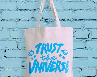 Peut inclure: Un sac fourre-tout en toile blanche avec du texte bleu qui dit "TRUST the UNIVERSE". Le texte est dans un style dessiné à la main et comprend un œil stylisé, une planète et des étoiles.
