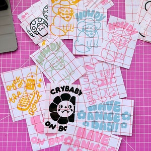 May include: A collection of colourful stickers featuring various designs, including a crying sunflower, a cat wearing a cowboy hat, and the phrase "Have a Nice Day!" The stickers are on a pink gridded surface.