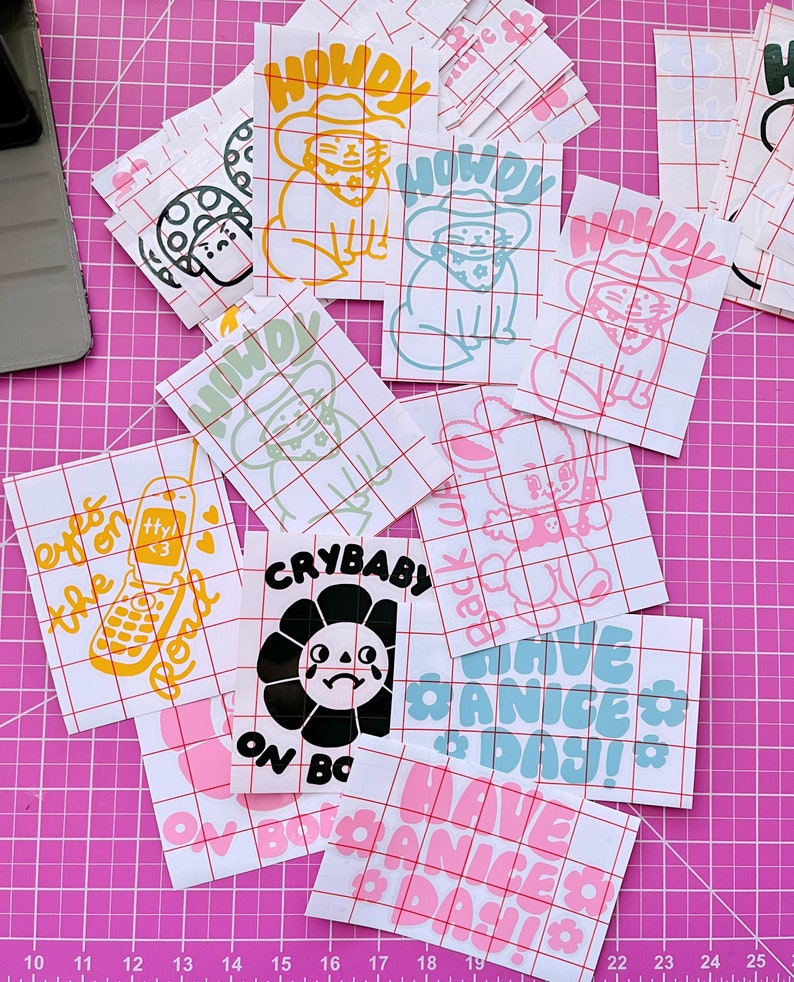 May include: A collection of colourful stickers featuring various designs, including a crying sunflower, a cat wearing a cowboy hat, and the phrase "Have a Nice Day!" The stickers are on a pink gridded surface.