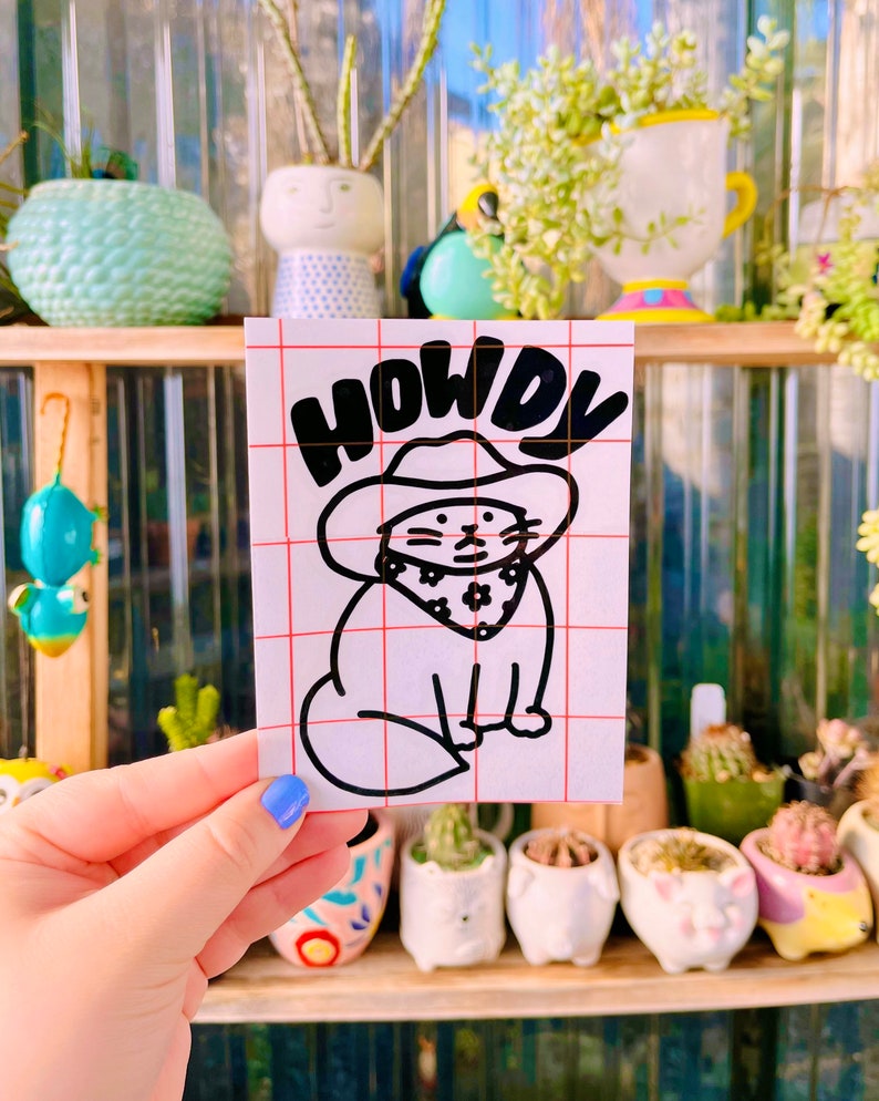 May include: A white sticker with a black outline of a cat wearing a cowboy hat and bandana. The text "HOWDY" is above the cat.