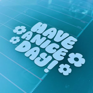 May include: A blue and white car window sticker with the text "HAVE A NICE DAY!" in a retro font with flowers on either side.