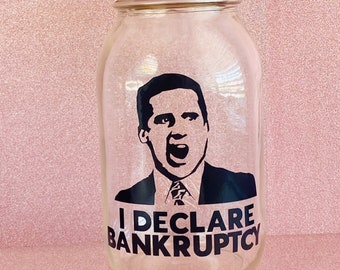 Peut inclure: Un bocal en verre transparent avec un couvercle doré. Le bocal est décoré d'une image en noir et blanc d'un homme avec le texte "I DECLARE BANKRUPTCY" imprimé dessus.