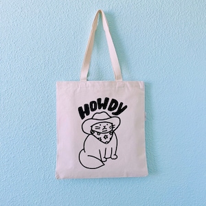 May include: A white tote bag with black text that says "HOWDY" and a cartoon cat wearing a cowboy hat and bandana.