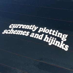 May include: White decal on a dark surface with the words "currently plotting schemes and hijinks" in a playful font. The decal is likely applied to a car window or similar surface.