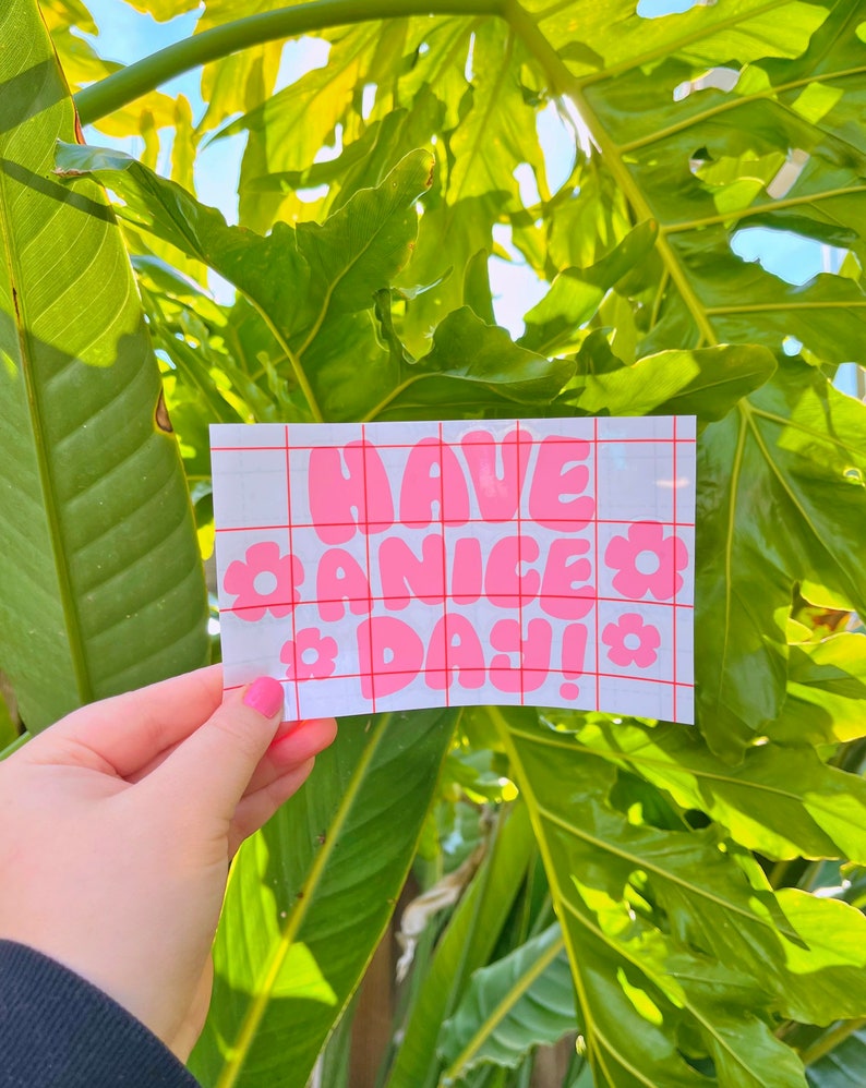 May include: A white sticker with a pink grid pattern and the words "HAVE A NICE DAY!" in pink bubble letters with pink flower accents.