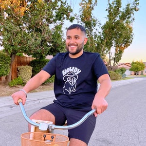May include: A person wearing a black t-shirt with a white graphic of a cartoon dog wearing a cowboy hat and sunglasses. The text "Howdy" is above the dog. The person is riding a bicycle with a brown basket.