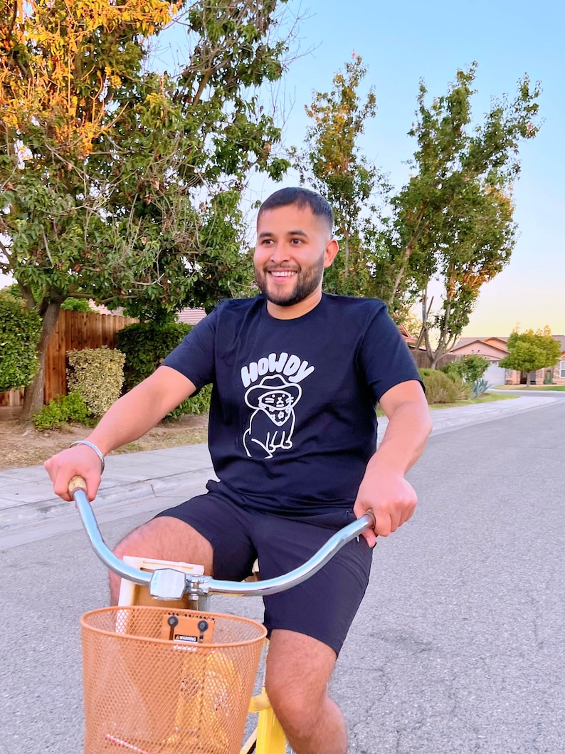 May include: A person wearing a black t-shirt with a white graphic of a cartoon dog wearing a cowboy hat and sunglasses. The text "Howdy" is above the dog. The person is riding a bicycle with a brown basket.