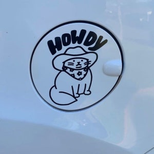 May include: A white car gas cap with a black and white sticker of a cat wearing a cowboy hat and bandana. The sticker says "Howdy" above the cat.