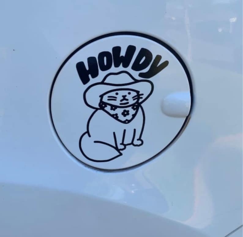 May include: A white car gas cap with a black and white sticker of a cat wearing a cowboy hat and bandana. The sticker says "Howdy" above the cat.