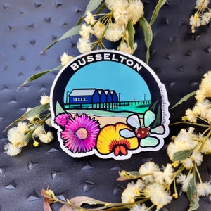 Może przedstawiać: Okrągła naklejka z niebiesko-białą ilustracją budynku z tekstem "Busselton" w czarnym kółku. Naklejka przedstawia błękitne niebo i plażę z kwiatami.