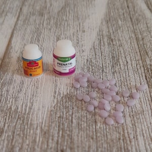 May include: Two small pill bottles, one labeled "Folic Acid" and the other "Prenatal", sit on a light wood-grain surface. Scattered around the bottles are numerous small, light purple pills. The bottles have white lids and colorful labels.