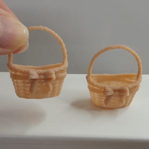 May include: Two miniature woven baskets with handles and bow details. One basket is held by a hand, the other sits on a small white table. The table is next to a white chair. The baskets are a light tan color.