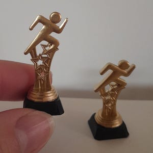 May include: Two small gold-colored running figure trophies. The trophies have a black base and a gold star design. One trophy is held by a person's hand.