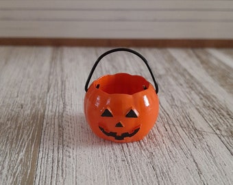May include: A small orange pumpkin-shaped trick-or-treat bucket with a black handle. The bucket has a carved jack-o'-lantern face.