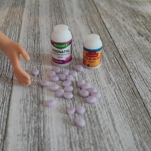 May include: Two small pill bottles labeled "PRENATAL" and "FOLIC ACID" sit on a wooden surface, with several small, light purple pills scattered around them. The bottles have white lids and colorful labels. A doll's arm is in the frame.