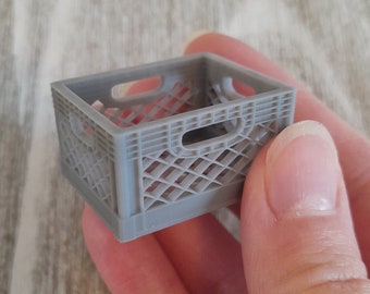 May include: A small, gray miniature crate with a lattice design. The crate has rectangular openings on the sides and a handle opening on each end. The crate is held in a person's hand.