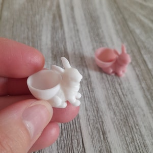 May include: A white bunny bowl is held in a hand, with a pink bunny bowl in the background. The bunny bowls are small, with a bowl-shaped cavity on the bunny's back. Text at the top says "1 bunny bowl included: either pink or white."