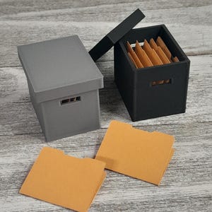 May include: A set of three ceramic mugs with a speckled, matte finish. The mugs are in shades of cream, light gray, and dark gray. Each mug has a simple, minimalist design and a rounded handle. The mugs are likely for coffee or tea.