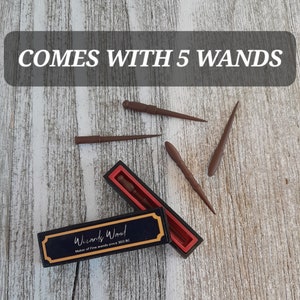 May include: The image shows five brown wands, some scattered and some in a black and red box. The box has a gold border and the text "Wizards Wand, Maker of Fine wands since 300 BC". The text "COMES WITH 5 WANDS" is at the top.
