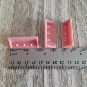 May include: Three pink dollhouse shelves, each with three holes, measuring approximately 1 inch in length. The shelves are shown on a wooden surface with a ruler for scale. Listing is for 1 dollhouse shelf.