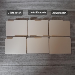 Op de afbeelding: Zes beige ordners met gekerfde lipjes, gerangschikt in twee rijen van drie. De bovenste rij heeft labels met de tekst "2 left notch", "2 middle notch" en "2 right notch". De ordners staan op een grijs houten oppervlak.