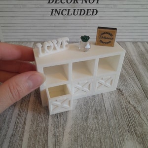 Peut inclure: Étagère cubique blanche avec un tiroir et trois étagères ouvertes. L'étagère est décorée d'une petite plante en pot et d'une pancarte qui dit "Bienvenue". Le mot "LOVE" est écrit en lettres blanches sur l'étagère supérieure.