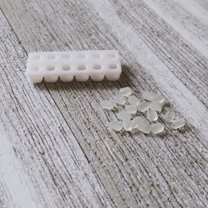 Può includere: Uno stampo di plastica bianca con 16 piccole cavità rotonde, accanto a una dispersione di perline traslucide di forma ovale. Lo stampo è rettangolare con un motivo a griglia di fori. Le perline sono sparse su una superficie di legno grigio chiaro e bianco.