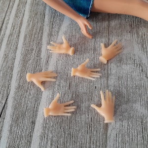 May include: Six small, pale pink doll hands are scattered on a light gray wooden surface. A doll's arm and denim shorts are visible in the background. The hands are in various positions, some open and some partially closed.