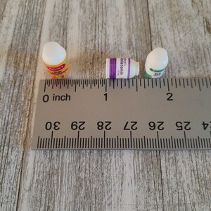 May include: Three small supplement bottles are arranged on a wooden surface next to a ruler. The bottles are labeled "Natural 1 Kids Multivitamin", "Melatonin", and "D3". The ruler shows measurements in inches.