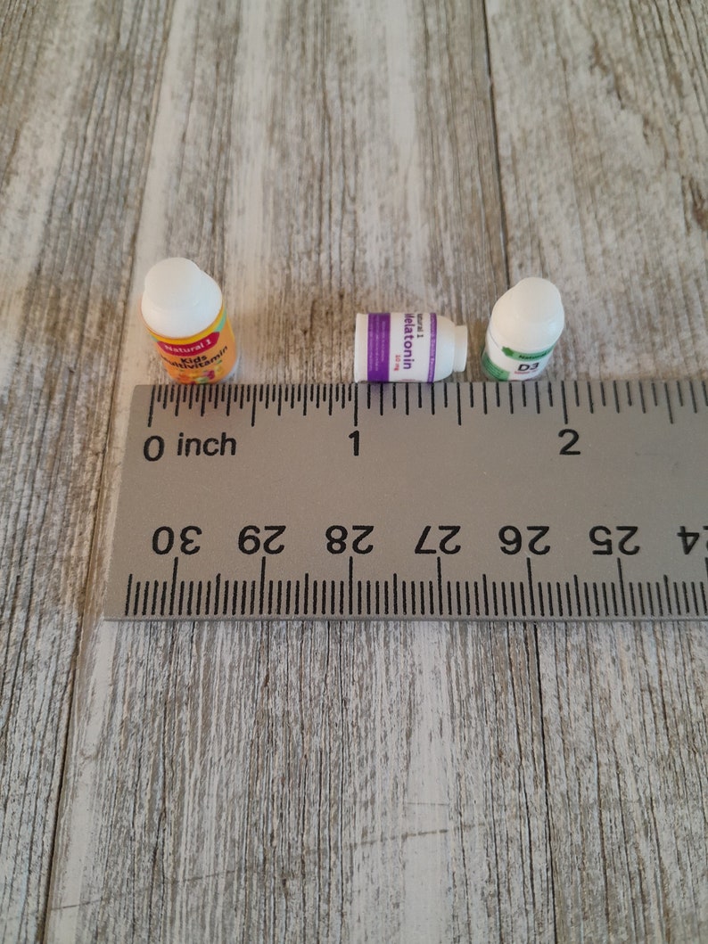 May include: Three small supplement bottles are arranged on a wooden surface next to a ruler. The bottles are labeled "Natural 1 Kids Multivitamin", "Melatonin", and "D3". The ruler shows measurements in inches.