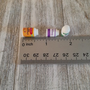 May include: Three small white supplement bottles with colorful labels are arranged near a ruler. The labels read "Kids Multivitamin", "Melatonin", and "D3". The ruler shows measurements in inches.