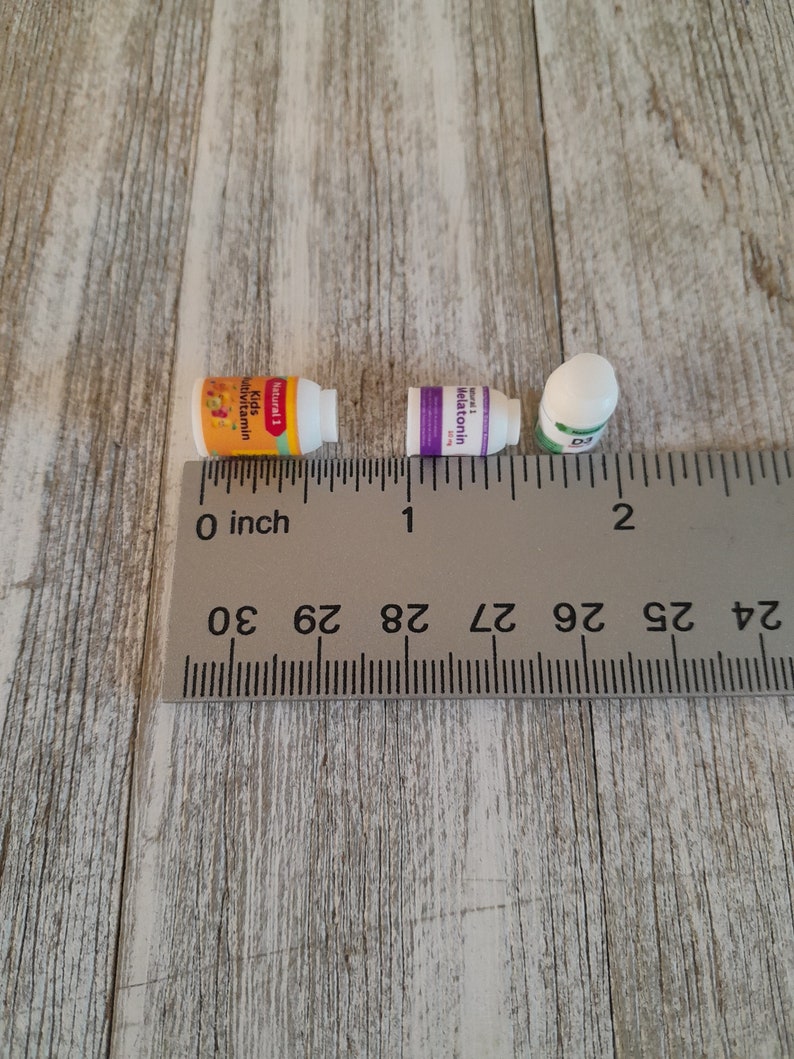 May include: Three small white supplement bottles with colorful labels are arranged near a ruler. The labels read "Kids Multivitamin", "Melatonin", and "D3". The ruler shows measurements in inches.