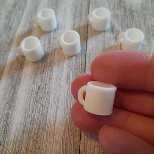 Pode incluir: Uma coleção de pequenas canecas em miniatura brancas. Uma caneca é segurada na mão, com as outras espalhadas sobre uma superfície clara com grão de madeira. Cada caneca tem uma pequena alça e formato cilíndrico, ideal para casas de bonecas.